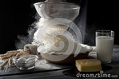 Flour sifting process in front of dark background. Glass of milk, eggs, cheese on wooden table. Stock Photo
