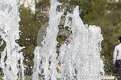 Flow splashes of water of city fountain, foam in jet of water. Fragment of recreation zone of the city park, green trees on Stock Photo