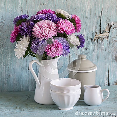 Flowers asters in a white enameled pitcher and vintage crockery - ceramic bowl and enameled jar, on a blue wooden background. Stock Photo