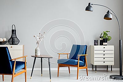 Flowers on wooden table between blue armchairs in grey flat interior with black lamp. Stock Photo