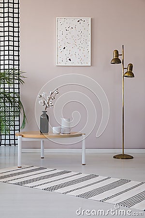 Flowers on wooden table next to gold lamp in living room interior with poster and rug. Real photo Stock Photo