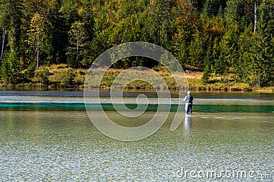 Fly fisherman stands in a waders in a lonely mountain lake while casting a fishing rod in the clear lonely lake in Bavaria in the Stock Photo