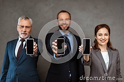 Focus of cheerful businessmen and businesswoman holding smartphones with blank screens on grey Stock Photo