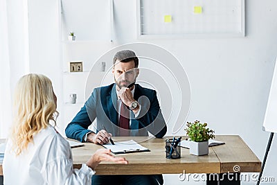 Focus of thoughtful recruiter in suit looking at blonde employee showing shrug gesture Stock Photo