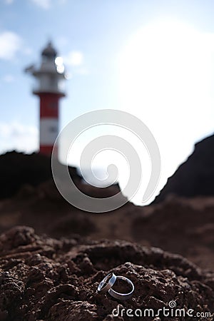 Focus on wedding rings on volcanic rock with blurred red and white lighthouse on the background Stock Photo