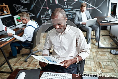 Envision more. Focused african male trader sitting by desk and studying analytical reports by looking through papers in Stock Photo