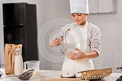 focused boy in chef hat and apron whisking eggs in bowl at table Stock Photo