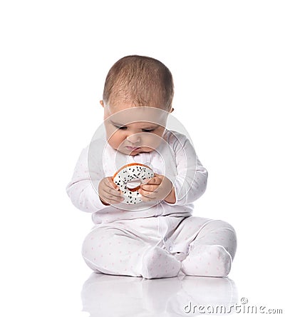 Focused infant baby toddler in white onepiece overall sits on the floor occupied with donut toy in hands Stock Photo