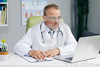 Focused middle aged senior head doctor in white medical coat and glasses sitting at workplace, talking to patient making Stock Photo