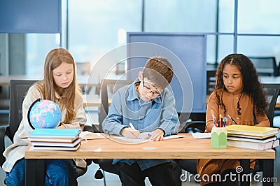 Focused multiracial students kids writing down data into notebook while sitting at table Stock Photo