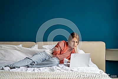 Focused teen girl working on study project on laptop lying in bed at home looking at screen. Stock Photo