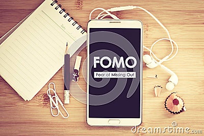 FOMO , Fear of missing out text on Smart phone with a Blank notepad and pencil on wooden table Stock Photo