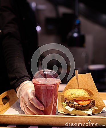 A tray with a burger on a foodcat. The chef& x27;s hand in a glove puts a glass with a drink on a tray. selective focus Stock Photo