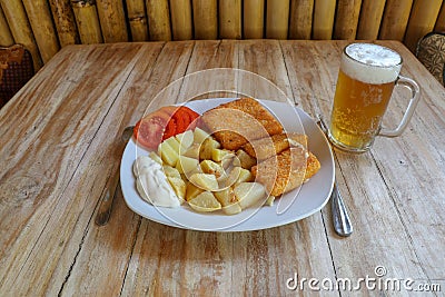 Food on white porcelain plate. Breaded fried cheese, boiled potatoes, tartar sauce, sliced tomato. Beer in glass on teak table. Stock Photo