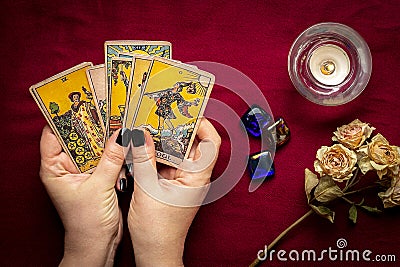 Fortuneteller`s hand with black manicure lays out tarot cards, crystal, candle, dry roses on red tablecloth Stock Photo