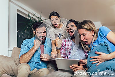 Four casual happy friends laughing shopping online together in a tablet sitting on a sofa in the living room Stock Photo