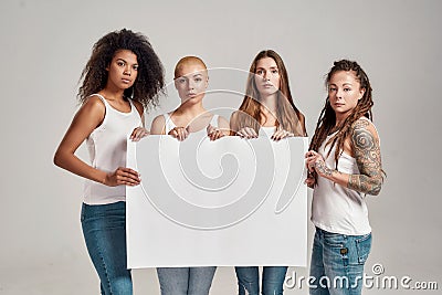 Four diverse women looking at camera while holding, standing with blank banner in their hands isolated over grey Stock Photo