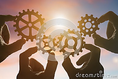 Four hands collect a puzzle of gears, against the background of the sky at sunset. Stock Photo