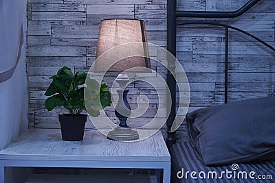 Fragment of a bedroom with a bedside table and a bed. A table lamp and a houseplant are on the nightstand Stock Photo