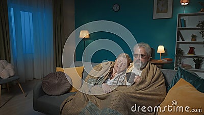 In the frame an elderly couple is sitting on a sofa under a plaid. The man stares intently into the distance with a cup Stock Photo