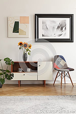 Framed photo and abstract art above sunflowers on an elegant cabinet and a patchwork chair in white living room interior. Stock Photo