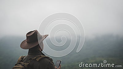 Freedom traveler man in hat carrying a backpack stands at the top of a mountain and using a smartphone on a foggy day.Adventure Stock Photo