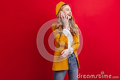 French woman in yellow jacket talking on phone. Studio shot of blonde girl in beret isolated on red background Stock Photo