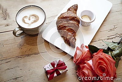 Fresh bakery croissant, coffee with heart sign, rose flowers on wooden table. Romantic breakfast for Valentine`s Day celebrate con Stock Photo