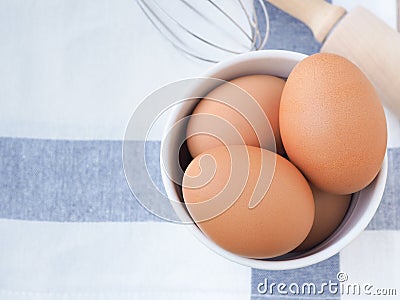 Fresh chicken eggs in a white bucket and whisk, rolling pin on a white dishcloth with blue straight stripes. Stock Photo