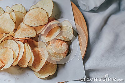 Fresh homemade deep fried crispy potato chips in white box on a wooden tray, top view. Salty crisps scattered on a table Stock Photo