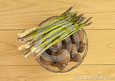 Fresh new potatoes with peel on and green asparagus in a glass bowl. Top view image on wooden background Stock Photo