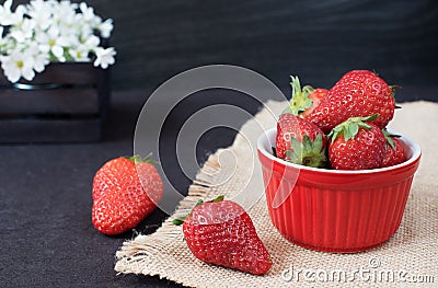 Fresh strawberries in mini red bowl on hessian jute. White and purple flowers in a decorative wooden crate. Black background Stock Photo