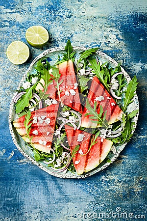 Fresh summer grilled watermelon salad with feta cheese, arugula, onions on blue background. Stock Photo