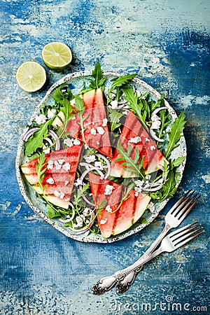 Fresh summer grilled watermelon salad with feta cheese, arugula, onions on blue background. Stock Photo