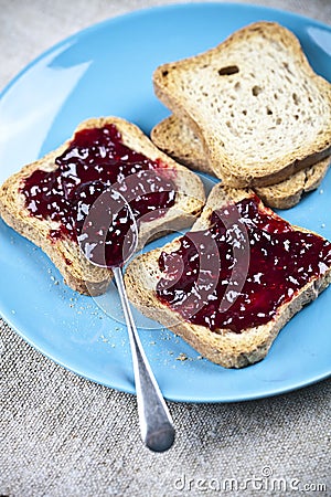 Fresh toasted cereal bread slices with homemade wild berries jam and spoon on blue ceramic plate closeup on linen tablecloth Stock Photo