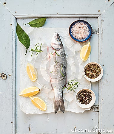 Fresh uncooked seabass fish with lemon, herbs, ice and spices on rustic blue wooden board backdrop Stock Photo