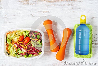 Fresh vegetable salad in lunch box with orange dumbbells excercise equipment and energy water drink on white rusty wood background Stock Photo
