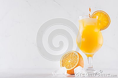 Fresh yellow oranges cocktail in elegance wineglass with ice cubes, straw and half oranges on soft white wood background. Stock Photo