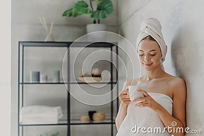 Fresh young European woman wrapped in towel after bath, stands in bathroom with mug of coffee or tea, enjoys morning, has pleased Stock Photo