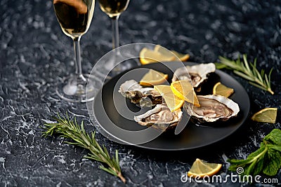 Freshly caught, opened oysters on a black plate with lemon slices, herbs and a glass of champagne on a dark stone background. Stock Photo
