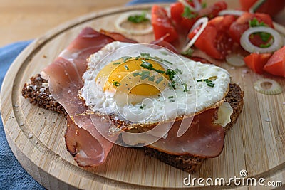 Fried egg and ham on a dark whole meal bread, tomatoes, onion rings and herbs garnish, served on a wooden cutting board Stock Photo