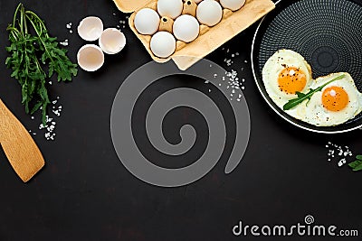 Fried eggs in frying pan with arugula leaves, salt and pepper for breakfast on black wooden background. Top view. Flat lay. Copy Stock Photo