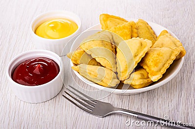 Fried savory pies in plate, bowls with mayonnaise and ketchup, fork on table Stock Photo
