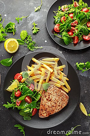 Fried Tuna Steaks on Black Plate with Fresh Green, Tomato Salad, lemon and french fries. healthy sea food Stock Photo