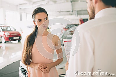 Friendly car salesman talking to a young woman and showing a new car inside showroom Signing of contract. Stock Photo