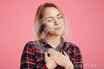 Friendly kind hearted female keeps eyes shut, hands on chest, expresses her gratitude to someone, poses against pink background. A Stock Photo