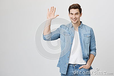 Friendly-looking attractive european student waives hand in hello gesture while smiling cheerfully and holding hand in Stock Photo