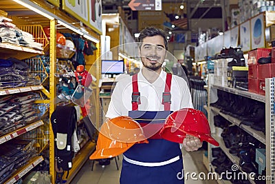 Friendly salesman demonstrates two colored safety helmets in industrial warehouse store. Stock Photo