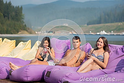 Friends with cocktails on cushioned loungers near the swimming pool on the background of river Stock Photo