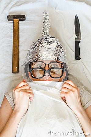 Frightened paranoid woman wears foil helmet and sleeps with weapon and different glasses because of mental illness Stock Photo